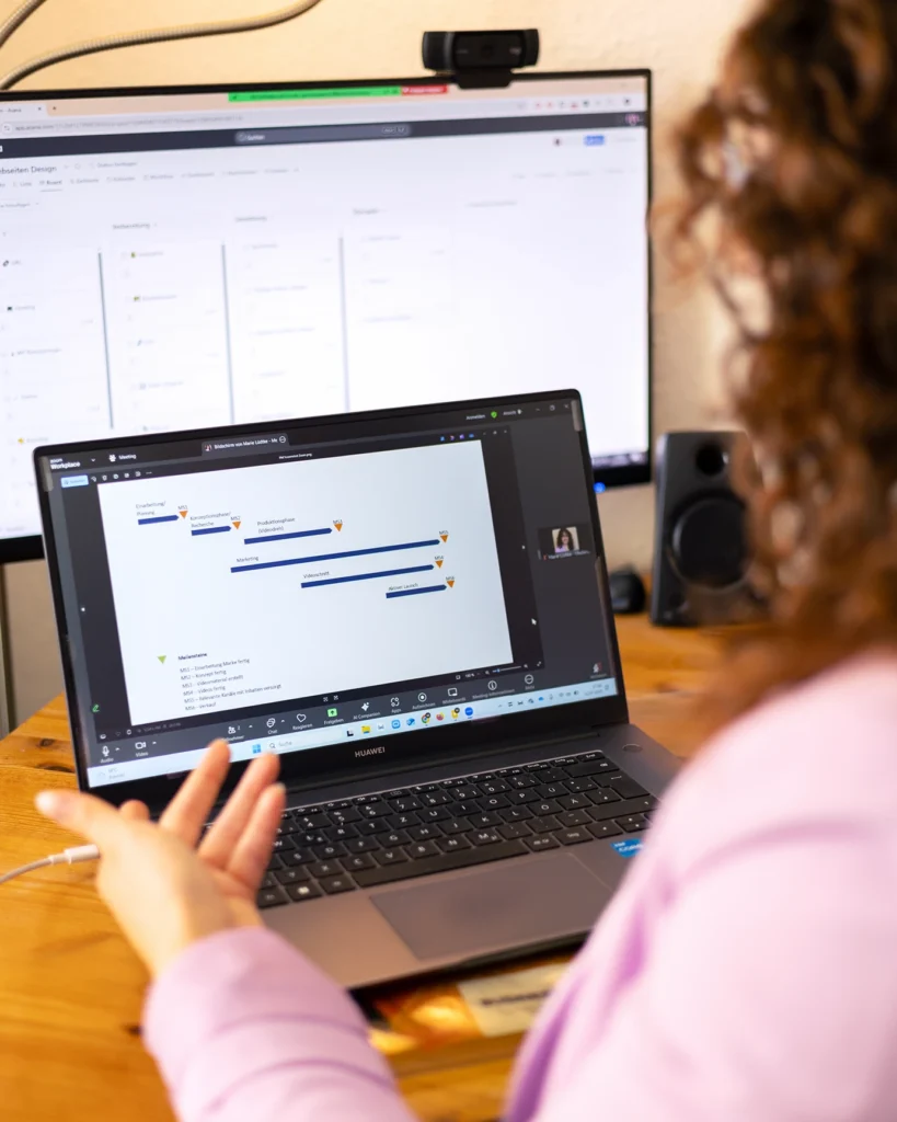 Sinnbild für Projektmanagement durch Marie Lüdtke, Gründerin von businessmarie: Blick über die Schulter von Marie Lüdtke, auf ihren Holzschreibtisch, auf welchem ein großer PC-Bildschirm und ein Laptop stehen. Beide sind angeschaltet und Marie Lüdtke hält ein Zoom-Meeting, indem sie über ein Projektmanagement spricht.