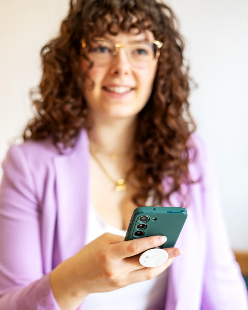 Sinnbild für strategische Launchbegleitung: Unscharfes Portrait von Marie Lüdtke, Gründerin von businessmarie. Im Hintergrund ist eine junge Frau mit langen, lockigen Haaren und Pony. Sie trägt ein weißes T-Shirt mit V-Ausschnitt, eine goldene Kette, eine goldene Brille und einen lila Blazer. Sie lacht freundlich an der Kamera vorbei. Im Fokus ist ihre Hand, die ein Smartphone hält.